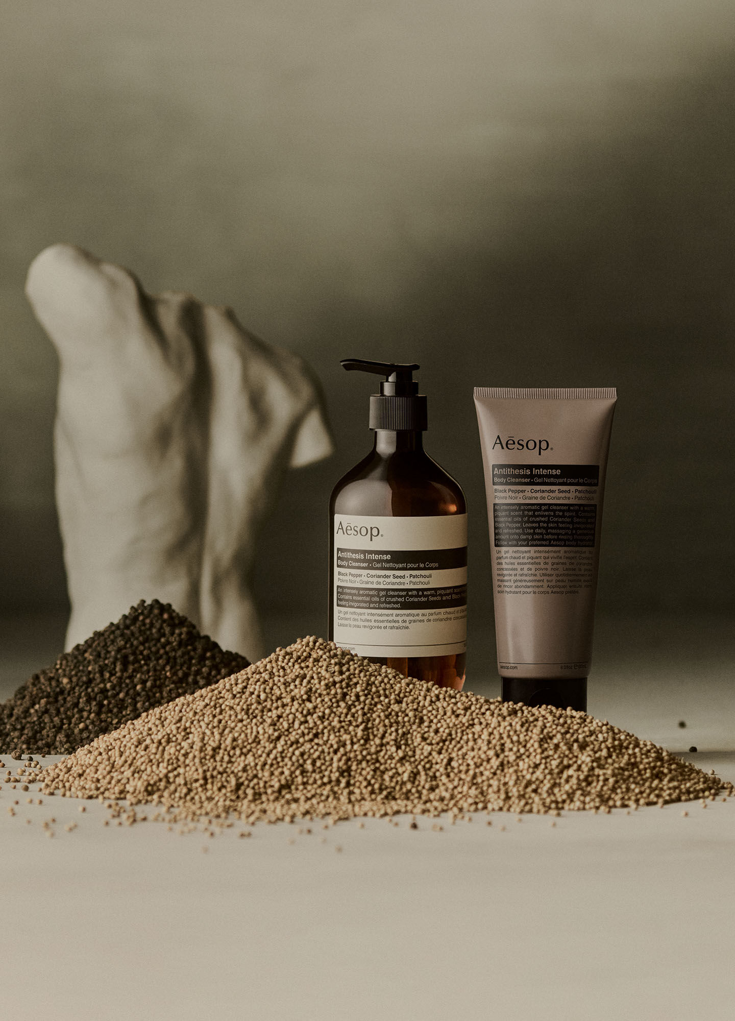 Aesop Antithesis Intense Body Cleanser pump bottle and tube beside piles of black peppercorns and coriander seeds.