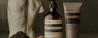 Aesop Antithesis Intense Body Cleanser pump bottle and tube beside piles of black peppercorns and coriander seeds.