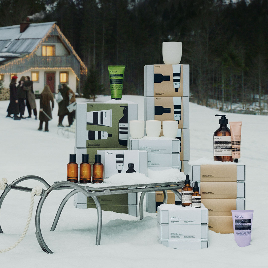 Aesop gift sets and products arranged on and around a sled in the snow, with a festively lit chalet and forest in the background.