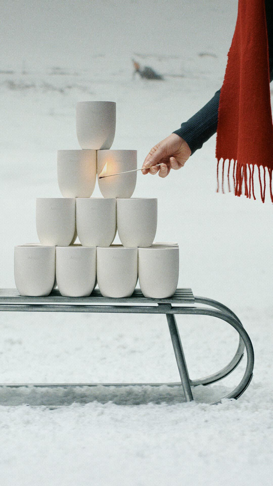 A hand holds a burning match beside white vessels stacked on a metal sledge in snow, with a red fringed scarf to the right.