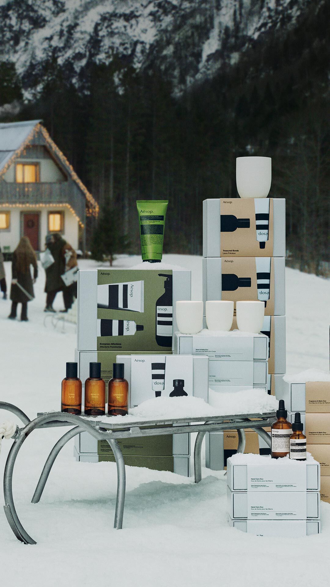 Aesop boxes, amber bottles, coloured tubes and white vessels are arranged around a metal sledge in snow, with people outside an illuminated house behind.